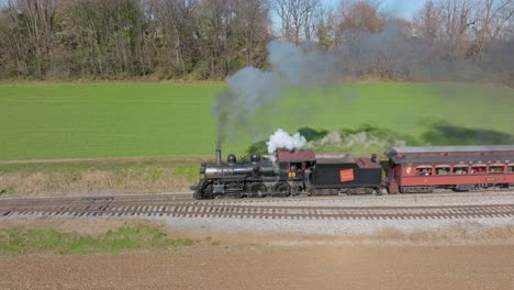 A-Drone-Side-View-of-a-Antique-Steam-Passenger-Train-Blowing-Smoke,-Traveling-Thru-the-Farmlands-on-a-Sunny-Fall-Day