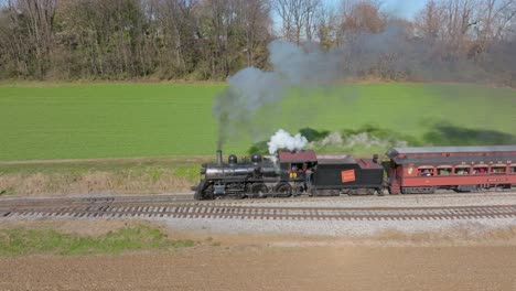 Eine-Drohnen-Seitenansicht-Eines-Antiken-Dampf-Personenzuges,-Der-Rauch-Bläst-Und-An-Einem-Sonnigen-Herbsttag-Durch-Das-Ackerland-Fährt