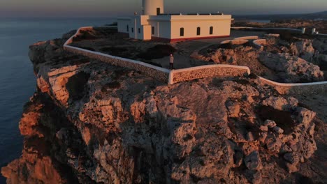 Cinematic-view-of-lighthouse-on-the-cliff-edge-seen-at-sunset-with-person-for-scale-standing-on-fence