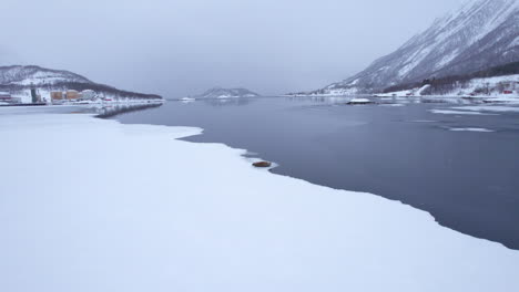 Slow-flyover-of-a-fjord-seal-laying-at-the-edge-of-sea-ice-in-a-fjord-during-a-light-snow-storm-with-snow-covered-mountains-and-factory-in-the-background