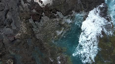Top-Down-View-Over-A-Volcanic-Rocky-Shoreline-With-Foaming-Waves-Splashing,-Sky-Reflecting-In-Small-Puddles