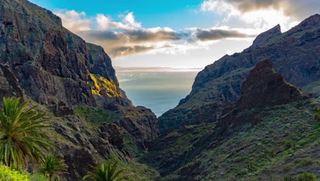 Sun-Setting-Over-Maska-Village-With-Palm-Trees-And-Green-Mountain-Peaks,-Orange-Sun-Rays,-Mountain-Valley,-Ocean-And-Blue-Sky-In-The-Background,-Time-Lapse,-Tenerife,-Spain