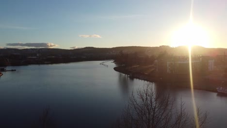 Sun-glaring-through-trees-during-ascending-aerial-from-Skien-Norway---Hjellevannet-lake-in-Telemark-canal-facing-north-towards-Stromdal-area