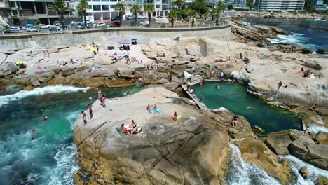 People-relaxing-on-boulders-at-Saunders-Rocks-Beach-in-Sea-Point,-Cape-Town,-aerial