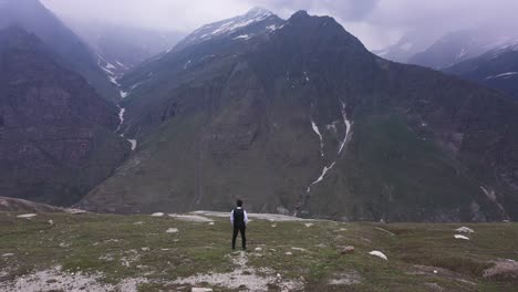 Aerial-shot-of-a-man-standing-in-the-edge-of-the-mountain-with-mountains-in-the-background