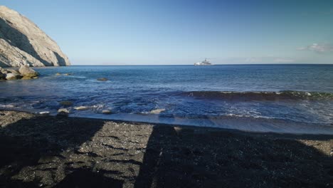 Panorama-Von-Entspannenden-Liegestühlen-Auf-Vulkanischem-Schwarzem-Sand-Im-Beach-Club-Mit-Blick-Auf-Die-Megayacht-Im-Meer-In-Santorini,-Griechenland