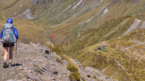 Static,-hiker-walks-exposed-mountain-trail,-distant-alpine-shelter,-Kepler-Track-New-Zealand