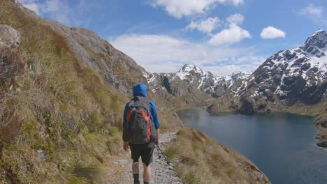 Follow,-hiker-walks-above-alpine-lake,-snow-capped-mountain-landscape,-Routeburn-Track-New-Zealand