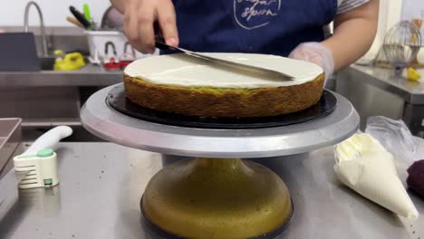 Front-view-of-a-professional-pastry-chef-smoothing-the-buttercream-meringue-frosting-with-spatula-while-rotating-the-revolving-cake-turntable-stand,-close-up-shot-in-commercial-bakery-setting
