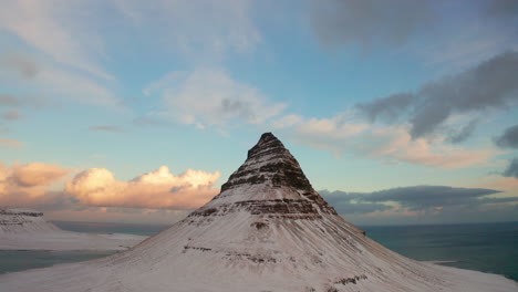 Kirkjufell-Mountain-Iceland-in-Winter,-Covered-with-Snow-Sharpened-Peak-at-the-Top-with-Long-High-Curved-Sides,-Aerial-Panoramic-Descending-View,-Blue-Cloudy-Sky-at-Sunset-in-Background