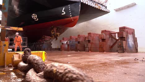 Ship-in-drydock-with-yard-workers-in-coveralls-walking-below-hull---Cinematic-low-angle-clip-with-movement-along-rusty-anchor-chain-in-foreground---Westcon-Norway