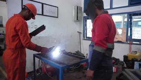 The-two-handymen-performing-welding-and-grinding-at-their-workplace-in-the-workshop,-while-the-sparks-"fly"-all-around-them,-they-wear-a-protective-helmet-and-equipment