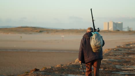 Lonely-old-fisherman-walking-on-a-sea-breakwater-at-sunset,-medium-shot