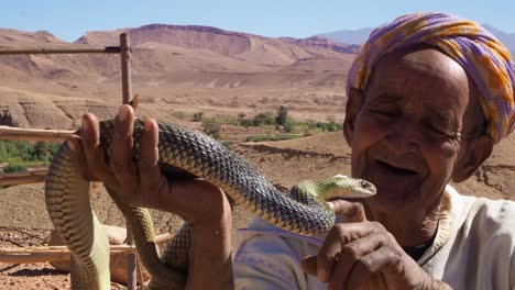 4K-Aufnahmen-Eines-Alten-Schlangenbeschwörers,-Der-Eine-Kobra-In-Der-Nähe-Der-Kasbah-Ait-Ben-Haddou-Im-Atlasgebirge-Von-Marokko-Hält