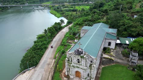 Stunning-Aerial-View-of-Vintage-Catholic-Church-facing-massive,-idyllic-river-with-lush-jungles-and-bridge-in-background