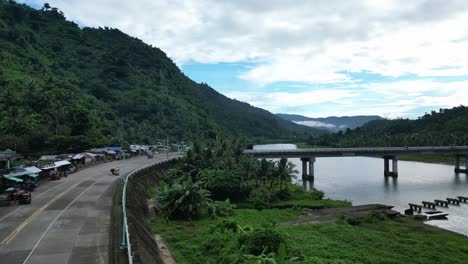 Rising-Overhead-Aerial-Shot-of-tricycles-and-vehicles-traveling-on-mountainside-roadway-leading-to-Bato-Bridge-amid-vast-river-and-lush-jungles