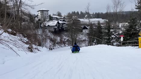 Downhill-Fahrer-Mit-Blau-schwarzer-Jacke-Fährt-Auf-Einem-Schlitten-Einen-Schneebedeckten-Hügel-Hinunter,-Mit-Kleinen-Häusern-Im-Hintergrund-Und-Einem-Winterwald-Am-Rande