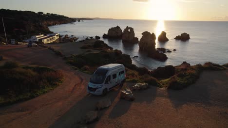 Aerial-establishing-view-campervan-parked-at-Praia-dos-Arrifes-glowing-sunlit-sunrise-Portugal-shoreline