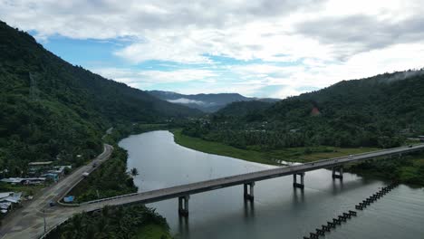 Establishing-Aerial-View-of-modern-bridge-over-tropical-river-with-breathtaking-cloudscape-and-lush-mountains-in-background