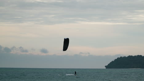 Man-kitesurfer-silhouette-while-kitesurfing-at-sunset-against-dramatic-cloudy-sky-by-the-island