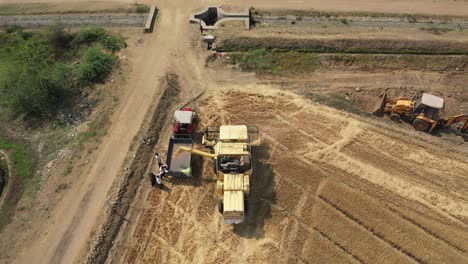 Un-Dron-Aéreo-Gira-Sobre-Una-Cosechadora-Agrícola-Que-Deja-Caer-Los-Granos-Cosechados-Recogidos-En-Un-Tractor-En-El-Campo-De-Trigo-Industrial-En-Un-Día-Soleado