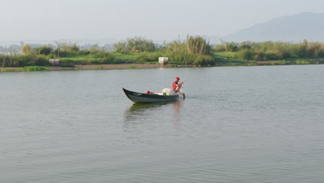 Tranquil-shot-of-southeast-asian-fisherman-adjusting-net-while-sitting-on-boat-in-middle-of-river