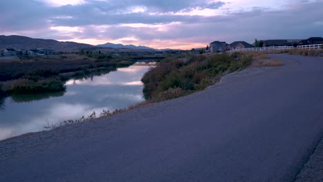 Person-riding-a-bike-along-a-trail-as-a-stunning-sunset-reflects-on-the-river---riding-away-from-static-camera