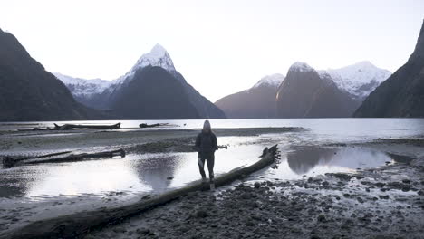 Slow-motion-shot-of-girl-in-hiking-gear-with-camera-walking-along-Milford-Sound-shoreline