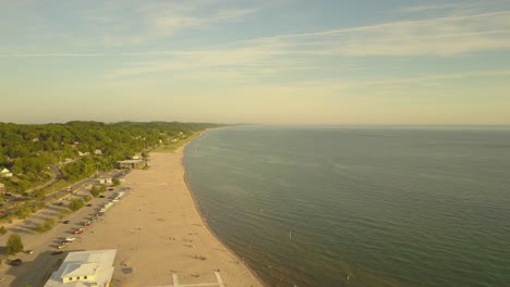 Drone-panning-shot-over-a-large-campground-beach-that-sits-along-the-lakeshore-with-blue-sky-and-blue-water-during-golden-hour-with-houses-in-the-background
