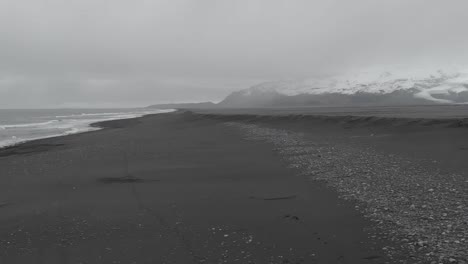 Aerial-shot-of-a-woman-running-along-a-black-sand-beach-with-panning-towards-the-mountains