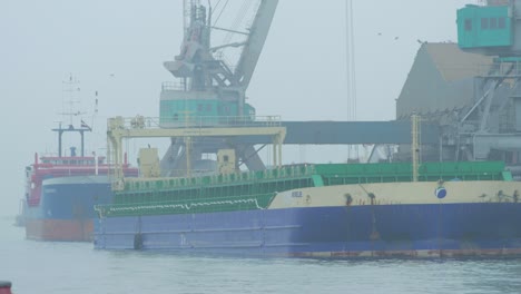 Port-cranes-loading-dry-cargo-ship-at-Port-of-Liepaja-in-foggy-day,-seagulls-in-frame,-wide-shot