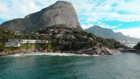 Aerial-panning-up-showing-the-beautiful-picturesque-cliff-rocks-of-Joatinga-beach-in-Rio-de-Janeiro-with-the-Gavea-mountain-towering-behind-it-and-the-green-ocean-waves-rolling-in