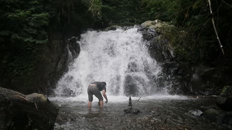 Hombre-Indonesio-Construyendo-Una-Torre-De-Roca-Equilibrada-De-Cairns-En-Un-Río-Frente-A-Una-Cascada