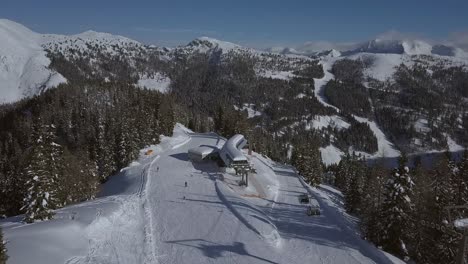 AERIAL-revealing-shot-of-a-ski-resort-on-a-sunny-day-with-people-on-the-ski-slope-and-in-the-ski-lift-with-beautiful-mountains-in-the-background