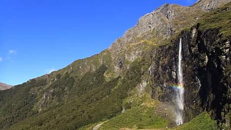 handheld-shot-of-the-waterfall-and-a-rainbow-near-the-Rob-Roy-glacier,-in-Wanaka,-New-zealand,-on-a-sunny-day