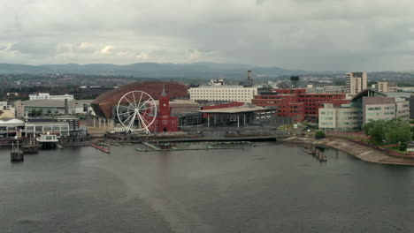 Narrow-Aerial-Flyover-of-Cardiff-Bay-Pulling-Back-from-the-Quayside-on-a-Cloudy-Summer’s-Day-with-Passenger-Ferry---Sailboat-in-Foreground