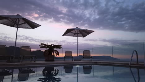 Sunset-Time-Lapse-of-Luxury-Greek-Villa-Patio-with-Pool,-Sun-Loungers---Umbrellas-in-Foreground-and-Mountains-in-the-Background
