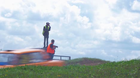 Autocross-cars-jumping-over-the-hill-in-amateur-racing-on-the-dirt-track-in-sunny-summer-day,-medium-low-angle-shot-with-beautiful-clouds-and-race-track-safety-man-and-photographer-in-background