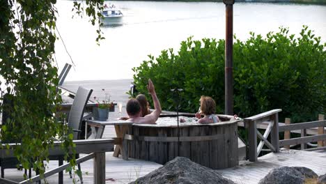 Girl-and-champagne-drinking-grandparents-in-outdoor-bathtub,-waving-at-approaching-boat-guests,-Nordic-summer-in-the-archipelago