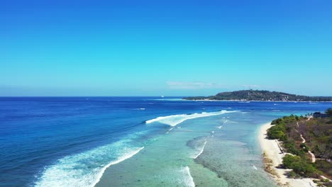 White-waves-of-blue-sea-foaming-over-coral-reefs-and-rocky-seashore-of-tropical-island-in-Indonesia,-with-a-bright-clear-blue-sky-background