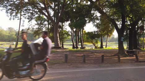 Toma-Estabilizada-De-Conducción-A-Través-De-Angkor-Wat-Con-El-Sol-Brillando-A-Través-De-Los-árboles-Al-Lado-De-La-Carretera