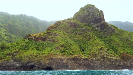 4K-Hawaii-Kauai-Boating-on-ocean-passing-cave-with-waves-crashing-on-rocky-shoreline-with-mountain-in-cloudy-distance-and-boat-spray-in-foreground