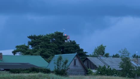 Fast-moving-dark-gray-rain-clouds-over-the-wooden-buildings-and-port-navigation-beacon-with-red-light-in-evening,-medium-shot