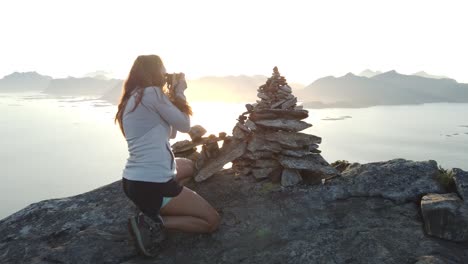 A-young-woman-with-long-hair-is-sitting-down-and-takes-a-photo-with-her-camera-of-a-small-pile-of-rocks-lying-on-top-of-each-other