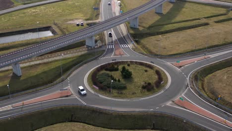 Aerial-wide-angle-fly-by-shot-of-a-white-car-on-a-highway-ring-road-roundabout-on-a-bright-sunny-afternoon