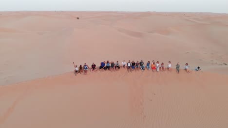 Drone-shot-of-friends-sitting-on-a-dune-and-waving-in-Abu-Dhabi-desert,-United-Arab-Emirates