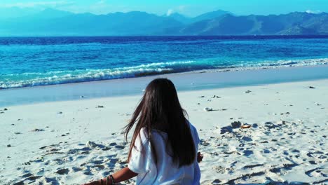 A-young-brunette-female-meditating-in-the-soft-warm-sand,-with-a-beautiful-view-of-the-mountains-in-the-distance