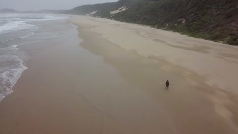 One-lone-fisherman-walks-along-wide-stretch-of-empty-beach,-aerial