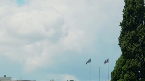 Griechische-Und-Europäische-Flaggen-Wehen-Neben-Einer-Zypresse,-Mit-Dem-Berühmten-Attika-Himmel-Im-Hintergrund