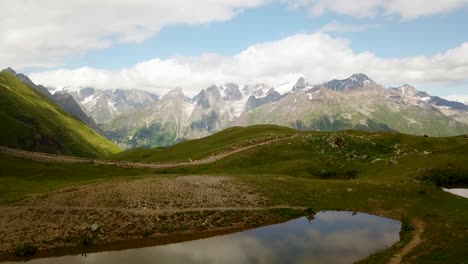 Wandern-Sie-Auf-Dem-Ruhigen-Koruldi-See-In-Mestia,-Einem-Grünen,-Frischen-Hochlandhügel-Mit-Einer-Wunderschönen,-Erstaunlichen-Landschaft-Der-Kaukasusberge-Mit-Schnee-Und-Blauem-Himmel-Mit-Wolken-Als-Spiegelbild-Der-Natur